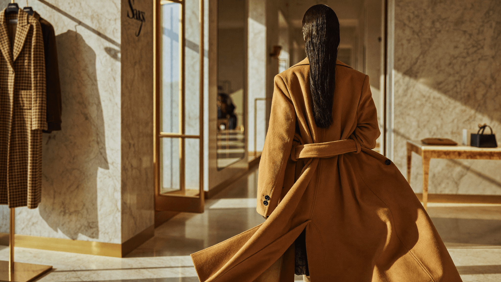 A woman in a camel wrap coat walking through a marble-and-brass atelier